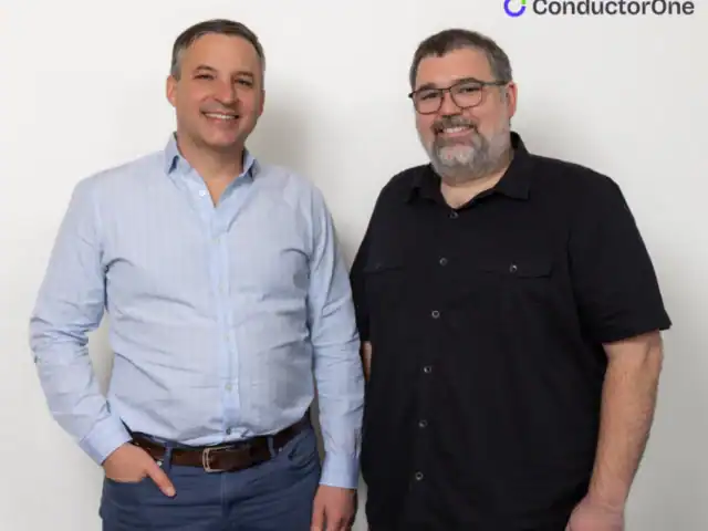Two men stand side by side and smile at the camera in front of a white wall with the ConductorOne logo in the upper right corner.